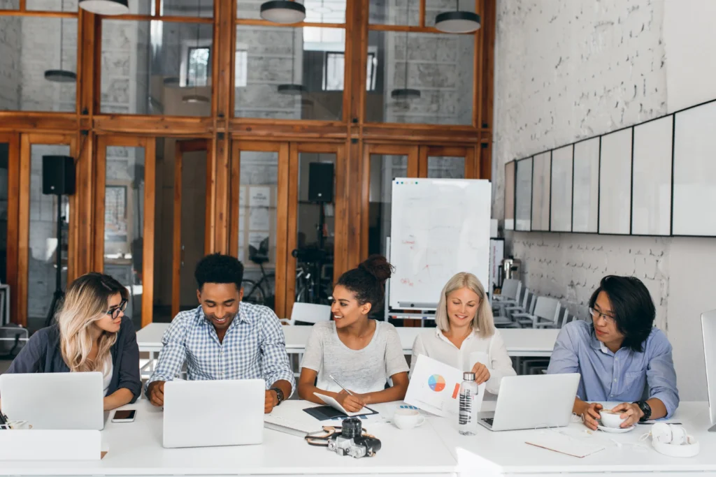 cinco pessoas de diferentes etnias e gêneros sentadas à mesa em um ambiente de trabalho com laptops e materiais impressos.